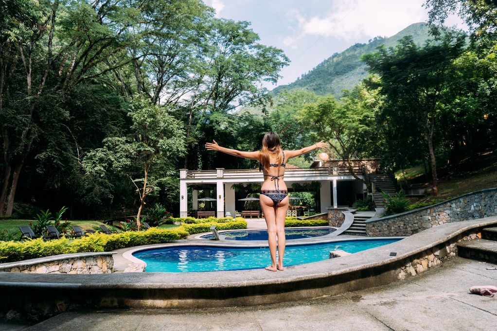 Woman in a bikini standing overlooking a hotel swimming pool with her arms apart